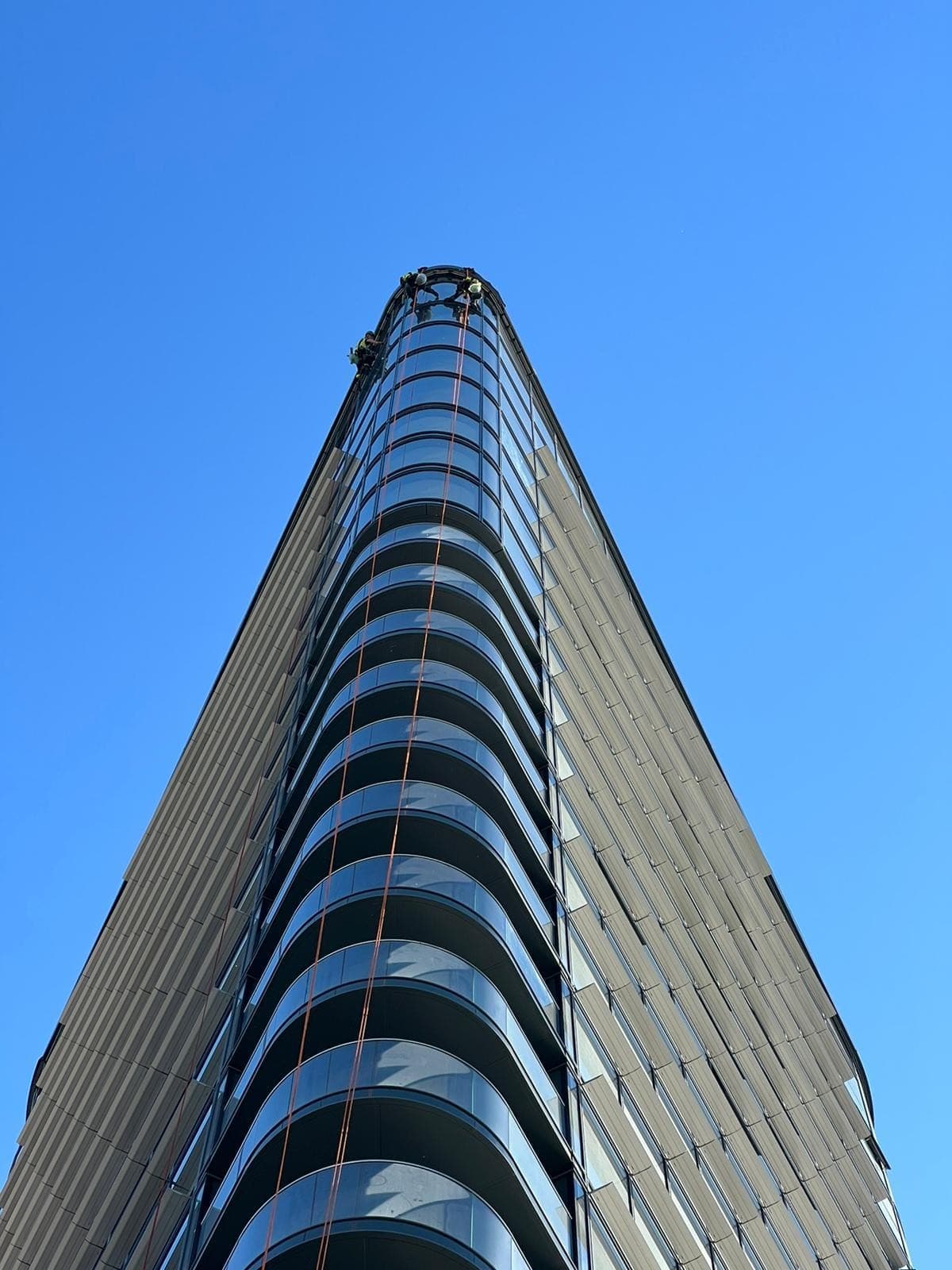 A tall glazed commercial tower photographed from street level, with rope access technicians working at the crown